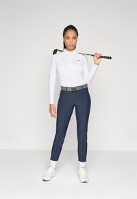 Femeie în picioare, ținând un golf club pe umăr, îmbrăcată cu un maiou alb cu mânecă lungă, pantaloni bleumarin, adidași albi și un pătraș gri.