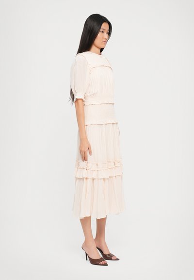 Nainen seisoo sivuttain päällä vaaleanbeige midi-mekko, jossa on röyhelöitä ja puffihihat, yhdistettynä ruskeisiin avonaisiin korkokenkiin.