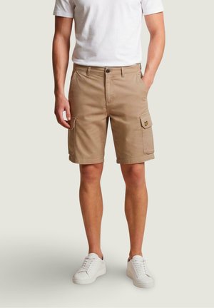 Mannequin homme portant un short cargo beige avec poches latérales et des baskets blanches, debout avec une main dans la poche devant un fond uni.