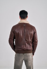 Hnědá kožená bunda s béžovým límcem z beránčí kožešiny, žebrovanými manžetami a lemem, hladkou texturou a viditelnými detaily stehů na zádech.