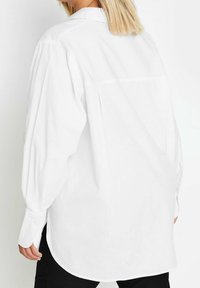 Person with shoulder-length blonde hair wearing a loose, long-sleeve white shirt with buttoned cuffs, shown from the back.