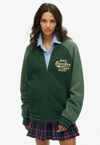 Veste bomber zippée verte avec une texture en zigzag, dotée d'un logo brodé NYC et de poches latérales, assortie à une chemise bleue à col et une jupe à carreaux.