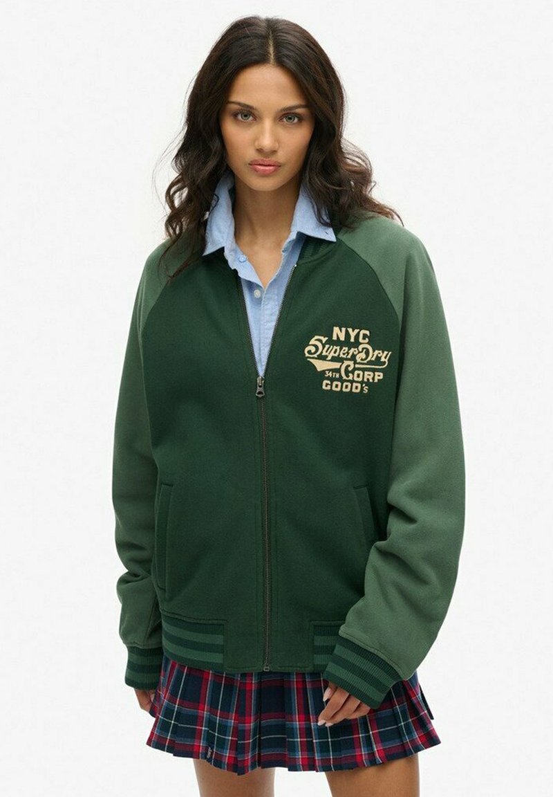 Veste bomber zippée verte avec une texture en zigzag, dotée d'un logo brodé NYC et de poches latérales, assortie à une chemise bleue à col et une jupe à carreaux.