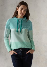 Capuchonpullover in gestreept licht- en donkerteal, met lange mouwen en trekkoorden. Draagt met donkere spijkerjeans, handen in de zakken.
