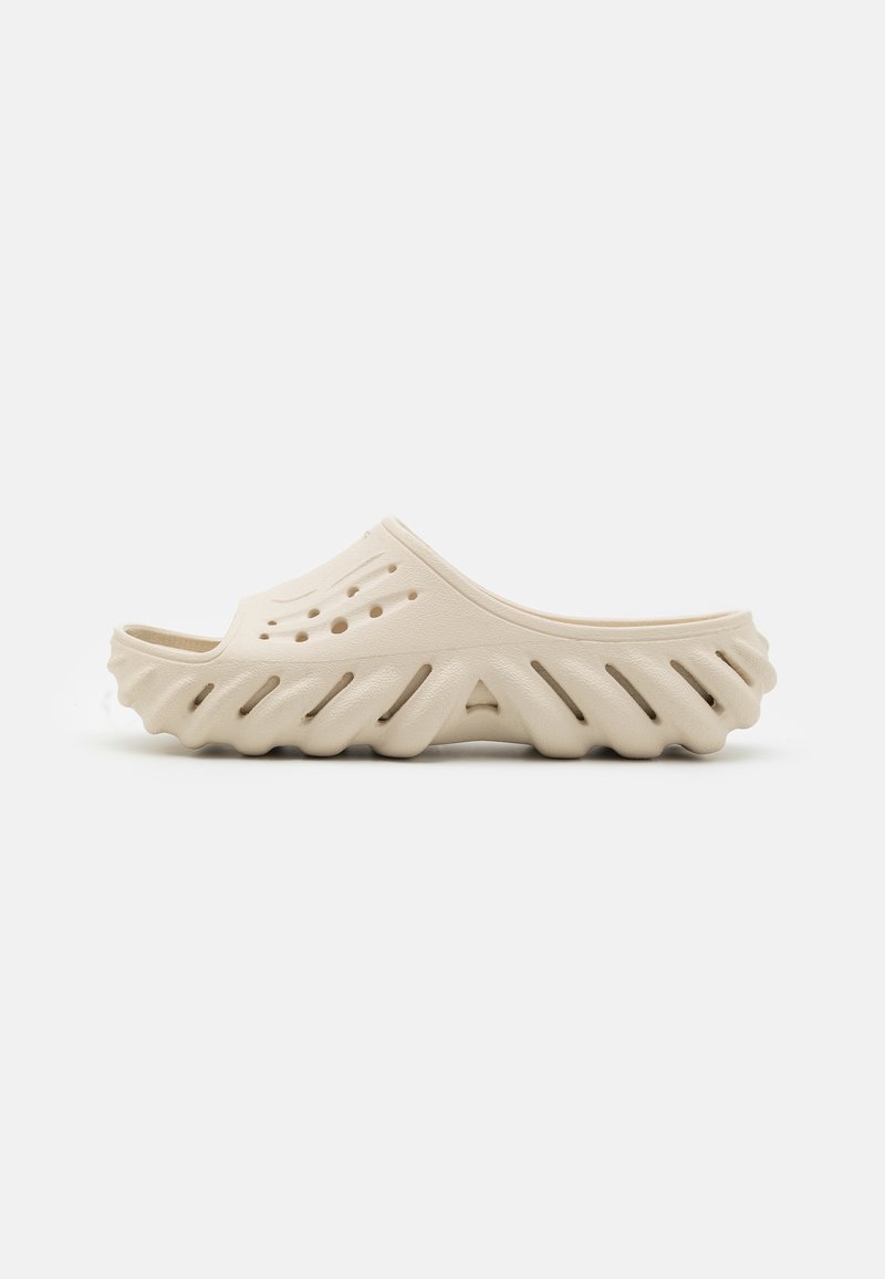 Sandale beige avec une semelle texturée et ondulée, dotée de trous de ventilation et d'un dessus lisse. Matériau léger et design à arrière ouvert.