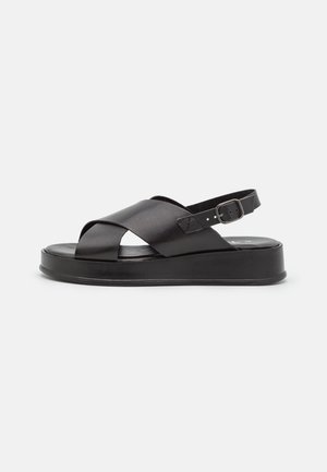 Sort platform-sandal med krydsede læderremme og justerbar hælrem med spænde på hvid baggrund.