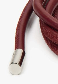 Červená kožená šňůra s hladkým, zaobleným kovovým zakončením; podél celé délky má pletenou texturu pro větší detail.