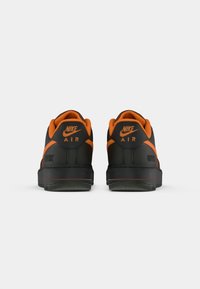 Nike Air tenisice s crnom gornjom stranicom i narančastim detaljima te teksturiranom potplatom. Na peti se nalazi Nike logo i naziv modela.