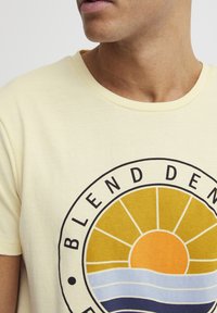 Kortermet gul bomull t-skjorte med en rund grafisk design av en sol og bølger i oransje, gule og blå nyanser.