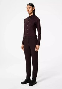 Femme se tenant debout dans une chemise boutonnée à motifs sombres, un pantalon sombre assorti et des bottes noires à la cheville, sur fond blanc.