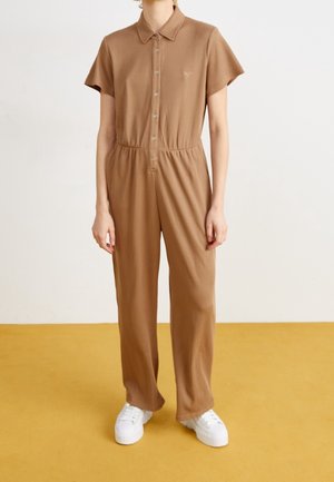 Une salopette marron à manches courtes avec un col et une fermeture à boutons, taille élastique et jambes larges, assortie à des baskets compensées blanches.