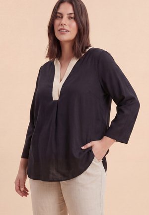 Vrouw die een zwart langarmig blouse draagt met een witte V-hals, gecombineerd met licht beige broek, staand met één hand in de zak.