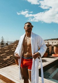 Vit badrock, röda shorts med svart midjeband, solglasögon; står på en träterrass med tak av kakel och pool i bakgrunden, blå himmel.