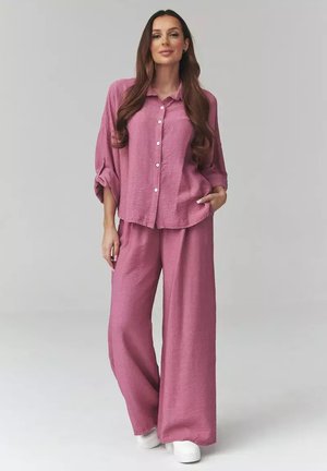 Kobieta stojąca pewnie, ubrana w luźną różową koszulę na guziki oraz pasujące szerokie spodnie i białe buty na jasnym tle.