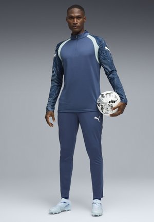 Mann in blauer Puma-Sportkleidung hält einen weiß-schwarz gemusterten Fußball und steht vor einem grau verlaufenden Hintergrund.