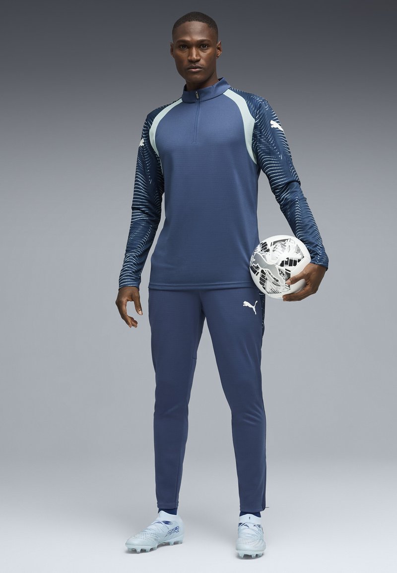 Mand iført blå Puma sportstøj, der holder en hvid og sort mønstret fodbold, stående mod en grå gradient baggrund.