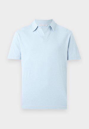 Jeune homme aux cheveux bouclés portant un polo bleu clair, un jean large en denim clair avec ceinture noire et des chaussures noires, debout avec une main derrière le dos.