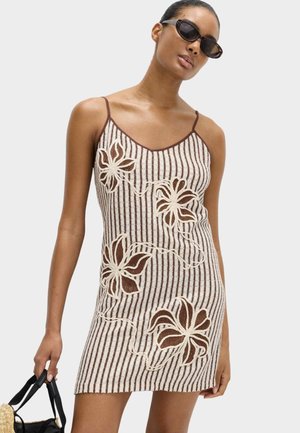 Kobieta w brązowo-białej sukience w paski z kwiatowym wzorem, w okularach przeciwsłonecznych cat-eye, trzymająca czarną torebkę ze słomy.