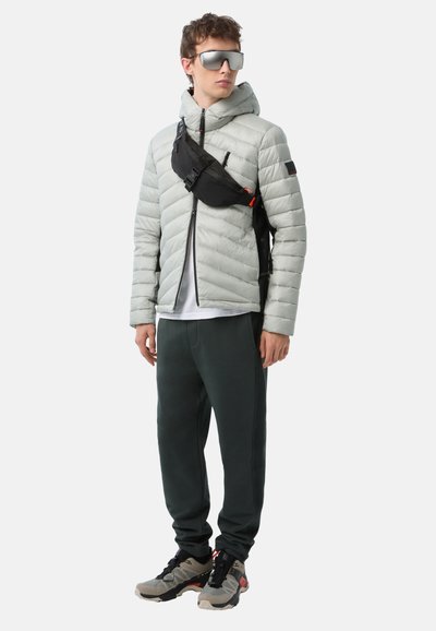 Hellgraue Steppjacke mit Kapuze, schwarze Brusttasche, weißes T-Shirt, dunkelgrüne Hose und klobige grau-schwarze Sneaker. Silberne Sonnenbrille.