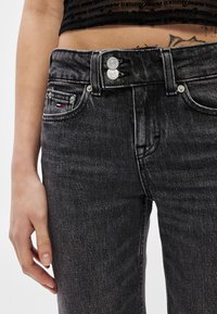 Čierne džínsové nohavice s vysokým pásom, vybavené dvojitým zapínaním na gombíky, prednými vreckami a malou štítkovou značkou na boku.