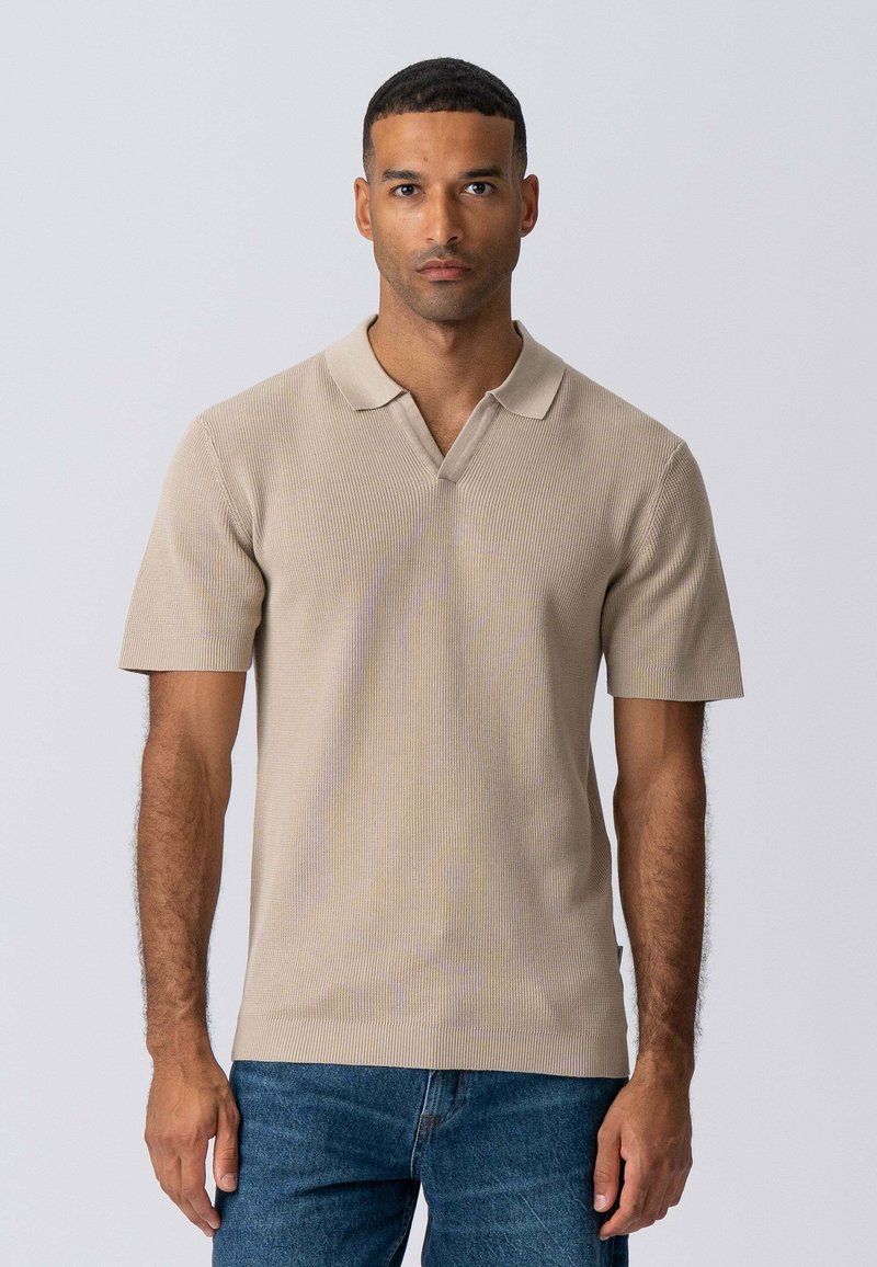 Beige ribbet polo shirt med V-udskæring og korte ærmer, lavet af tekstureret materiale. Sammen med blå jeans, præsenteret på en neutral baggrund.
