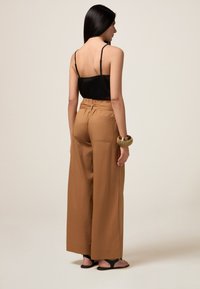 Camisola in satin nero e pantaloni ampi color cammello con vita arricciata, dotati di tasche laterali e un bracciale d'oro texture al polso.