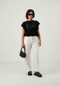 Blusa preta com folhos e detalhes em renda, combinada com calças brancas de perna larga. Acessórios incluem uma pequena mala preta e sandálias pretas chunky.