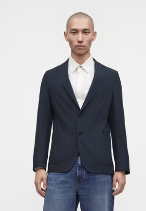 Uomo che indossa un blazer blu scuro sopra una camicia bianca e jeans blu, in piedi contro uno sfondo chiaro e uniforme.