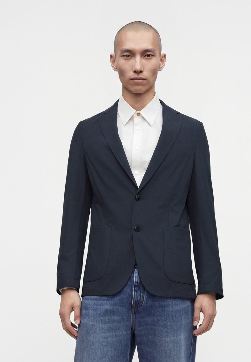 Homme portant un blazer bleu marine foncé sur une chemise blanche et un jean bleu, debout devant un fond clair uni.