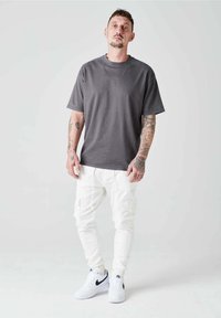 Camiseta gris de manga corta, pantalones cargo blancos y zapatillas blancas. Presenta tatuajes en los brazos y un diseño de ajuste relajado.