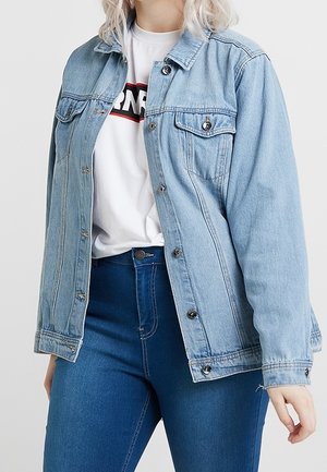 Femme portant une veste en denim bleu clair par-dessus un t-shirt graphique blanc et un jean taille haute bleu foncé, sur un fond uni.