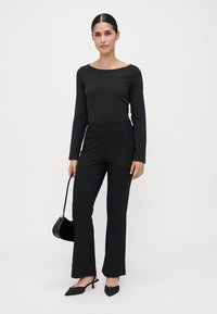 Woman in black long-sleeve top and pants holding black handbag, wearing black heeled shoes, standing against plain light background.