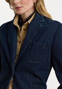 Donkerblauwe denim blazer met een gesneden kraag, een enkele voorkantzak en een gestructureerde afwerking, gedragen over een lichtbruin overhemd met knopen.
