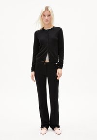 Svart stickad cardigan med knäppning, kombinerad med svarta byxor med fickor och en brun knäppning. Modellen bär ljusrosa ballerina.