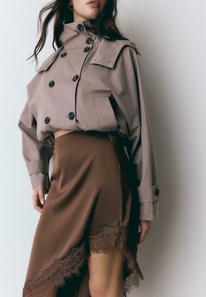 Frau trägt einen kurz geschnittenen beigen Trenchcoat mit großen Knöpfen und einen braunen asymmetrischen Rock mit Spitzenbesatz, steht vor einer weißen Wand.