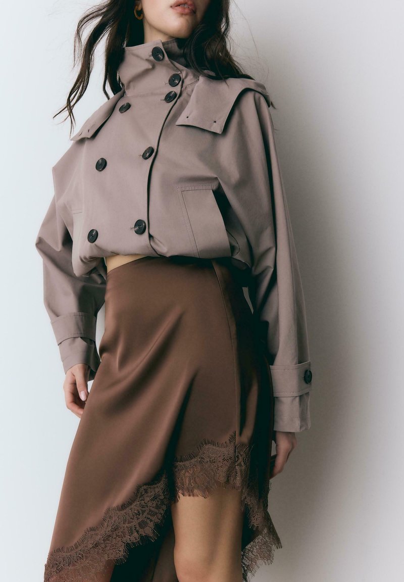 Frau trägt einen kurz geschnittenen beigen Trenchcoat mit großen Knöpfen und einen braunen asymmetrischen Rock mit Spitzenbesatz, steht vor einer weißen Wand.