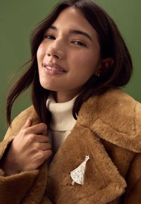 Faux fur brown coat, white ribbed turtleneck, silver tree brooch with star detail. Background is a solid green color.