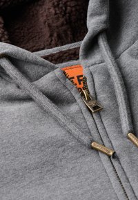 Grijze zip-up hoodie met een zachte, gestructureerde bruine fleece voering, een stevige messing rits en een oranje label met de tekst "SUPERDRY."