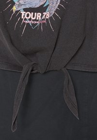 Černé tričko s uzlem na spodním lemu, zdobené barevným grafikou, která zahrnuje orla a text "TOUR 78" v růžové a modré barvě.