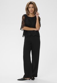 Top in pizzo nero con bordi scallopati, abbinato a pantaloni neri larghi e ballerine nere a punta, che mostrano un design a strati.