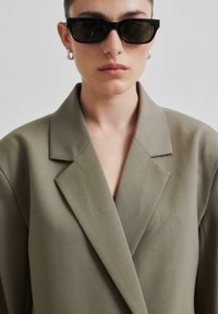 Överdimensionerad olivgrön blazer med strukturerade slag, slät tyg och v-ringning. Bärs med svarta rektangulära solglasögon och örhängen.