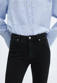 Jeans de mezclilla negra de cintura alta, con dos bolsillos frontales, un botón metálico y costuras en contraste, combinados con una camisa de botones azul clara.