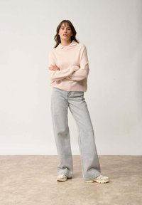Jasnoróżowy sweter z kołnierzykiem, o miękkiej fakturze, w połączeniu z jasnoszarymi, szerokimi spodniami z wysokim stanem oraz beżowymi trampkami, stojący na neutralnej podłodze.