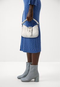 Femme portant une robe midi bleu côtelé et des bottines grises, tenant à ses côtés un sac à main blanc avec un détail de boucle.