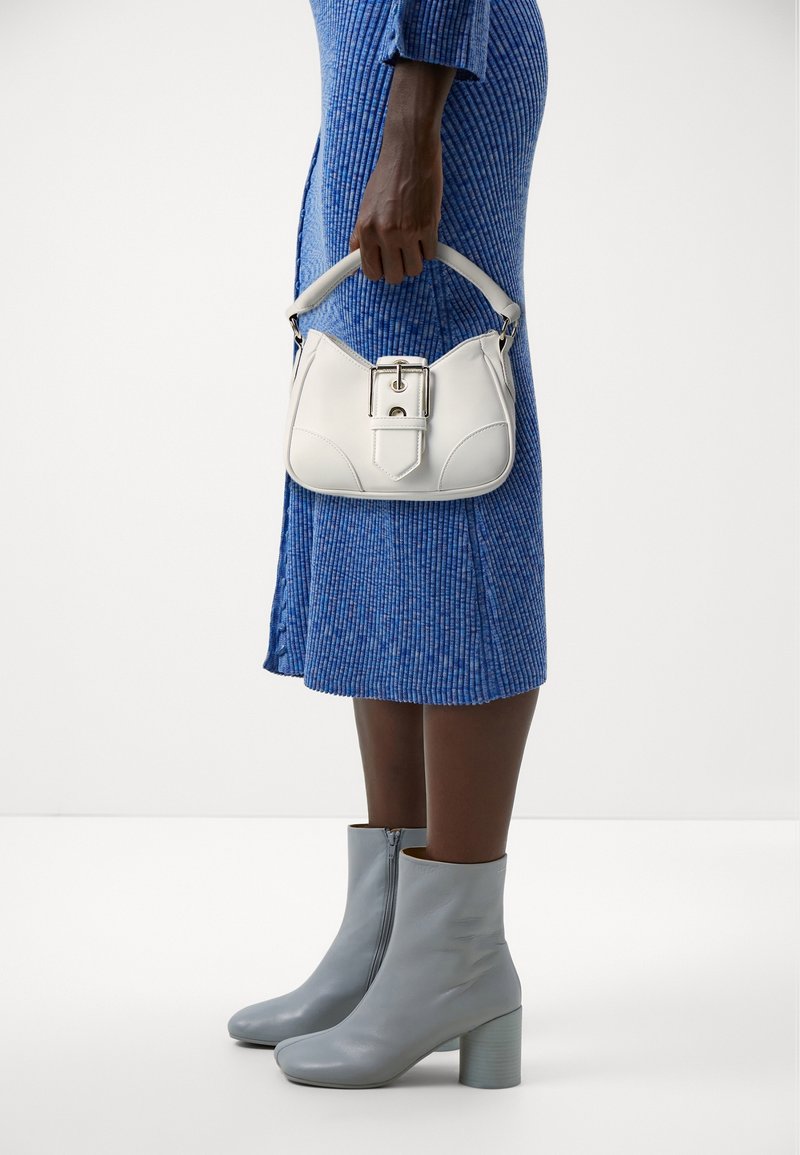 Femme portant une robe midi bleu côtelé et des bottines grises, tenant à ses côtés un sac à main blanc avec un détail de boucle.