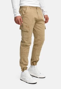 Homme portant un pantalon cargo beige avec poches latérales, des poignets élastiques, une chemise blanche à manches longues, des baskets montantes blanches et des chaussettes blanches, debout.
