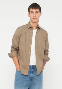 C camisa maro cu mâneci lungi, cu nasturi, cu un singur buzunar pe piept și mânecile suflecate, purtată peste un tricou alb și asociată cu blugi albaștri.