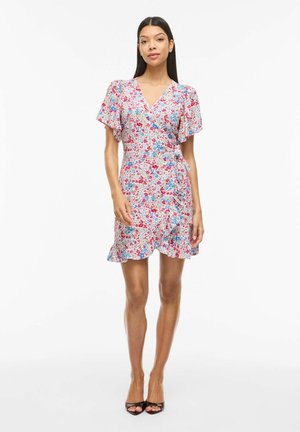 Femme debout portant une robe courte portefeuille fleurie avec un ourlet à volants et des manches courtes volantées, assortie à des talons ouverts noirs.