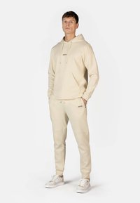 Beige hættebluse og matchende sweatpants, begge med en frontlomme og subtil branding. Letvægtsstof med en glat tekstur. Hvide sneakers.
