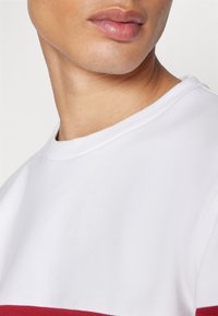 Hvid bomuld t-shirt med en rød horisontal stribe. Har rund halsudskæring og tekstureret stof. Nærbillede af kraven og skulderen.